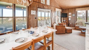 A dining area with a large table set for six, wooden chairs, and a view of the outdoors through large windows.