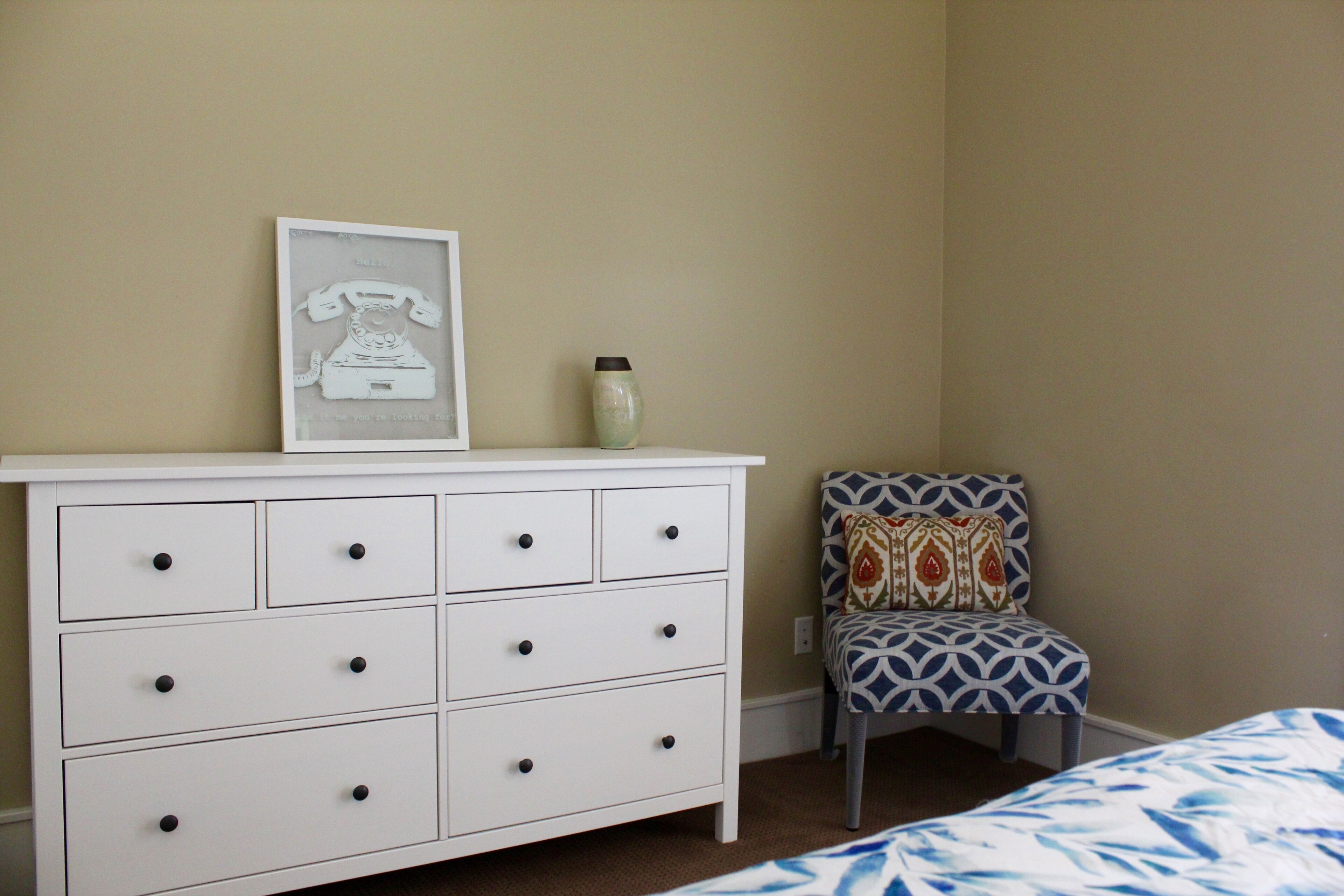 A bedroom with a white dresser, a patterned chair, and a bed with a blue and white patterned bedspread.