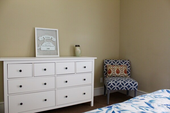 Un dormitorio con un aparador blanco, una silla estampada y una cama con colcha azul y blanca.