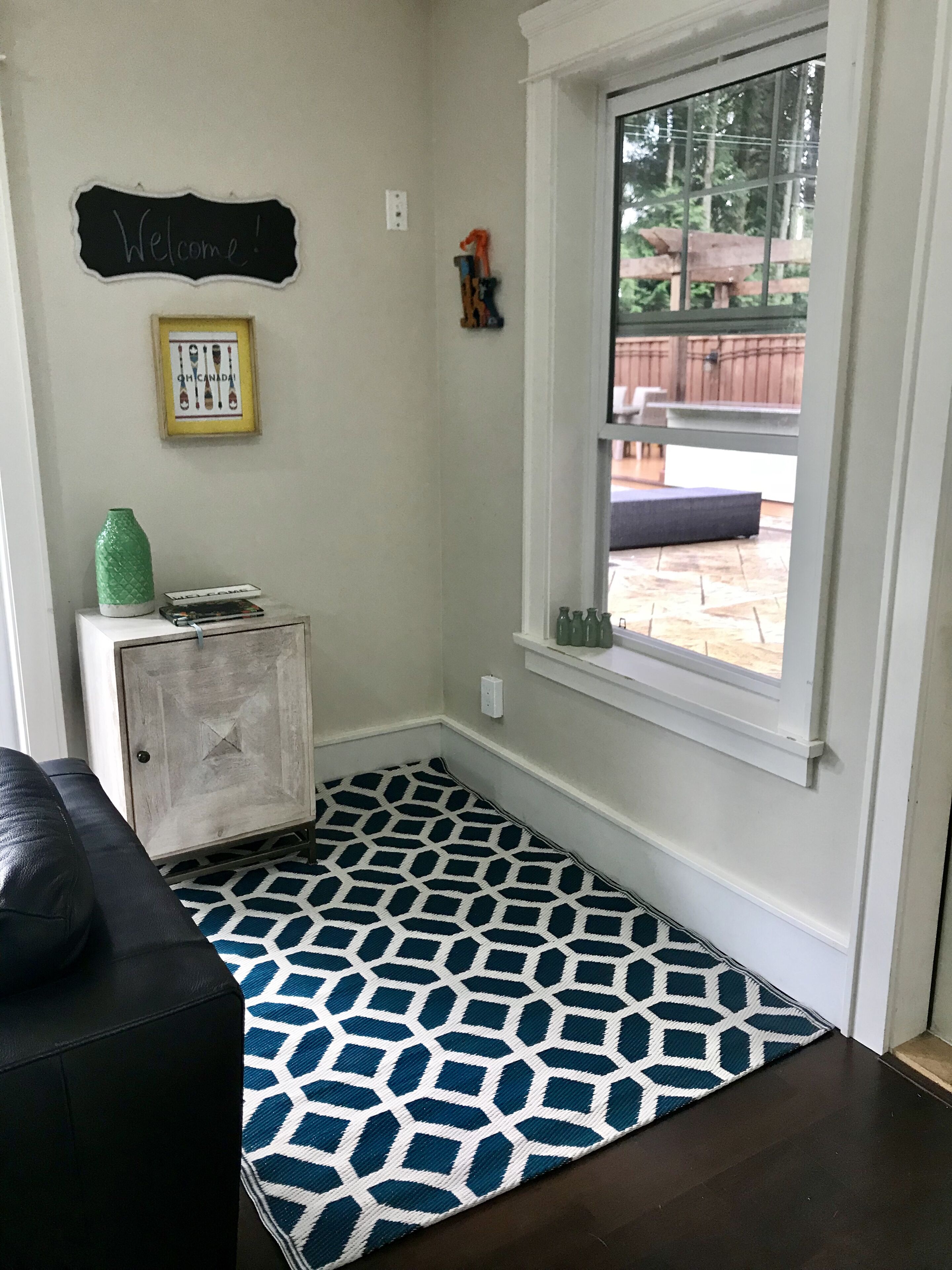 A room with a patterned rug, a small cabinet, a framed picture, and a window with a view of an outdoor patio.