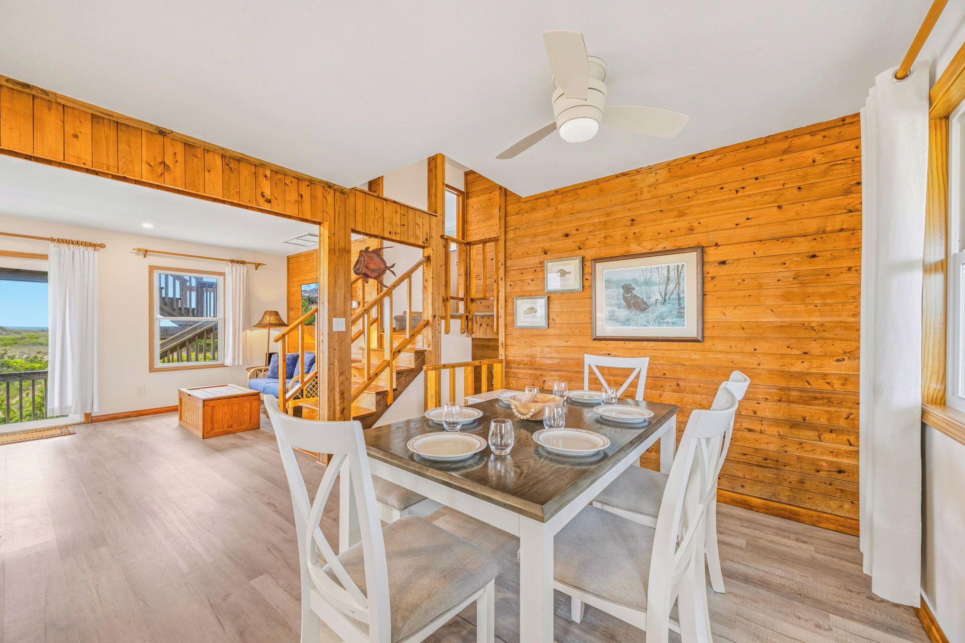 Un coin repas avec une table en bois dressée pour quatre personnes, des chaises blanches, un ventilateur de plafond et un escalier menant à un autre niveau.