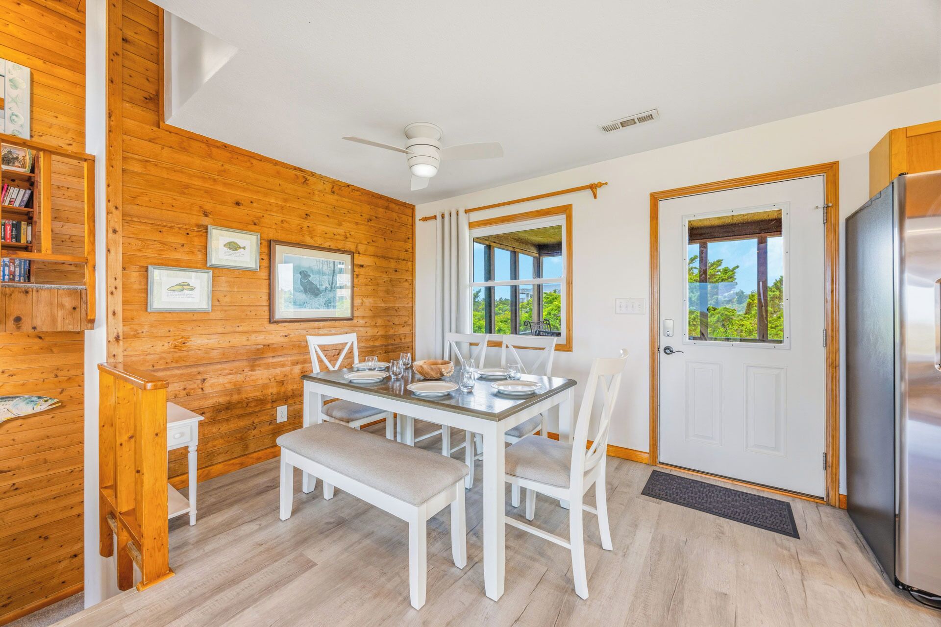 A wooden interior with a dining table set for four, a white door with a view, and a refrigerator.