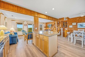 A spacious kitchen with wooden cabinets, a dining area with white chairs, and a view of the outdoors through large windows.