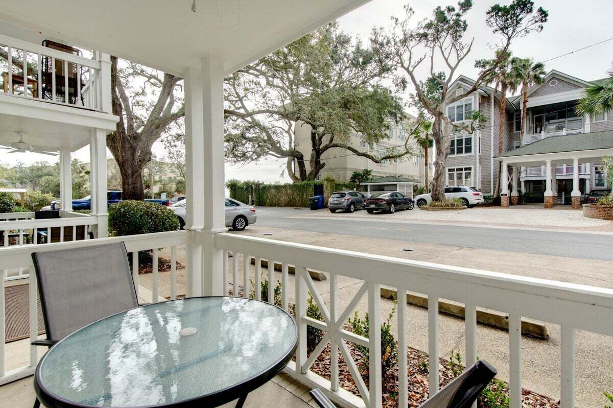 A covered outdoor seating area with a table and chairs, a white railing, and a view of a street with parked cars and a large tree.