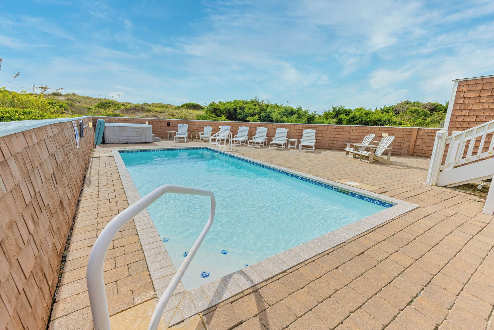 A rectangular outdoor swimming pool with a tiled edge, surrounded by a brick deck. There are white lounge chairs and a hot tub on the deck.