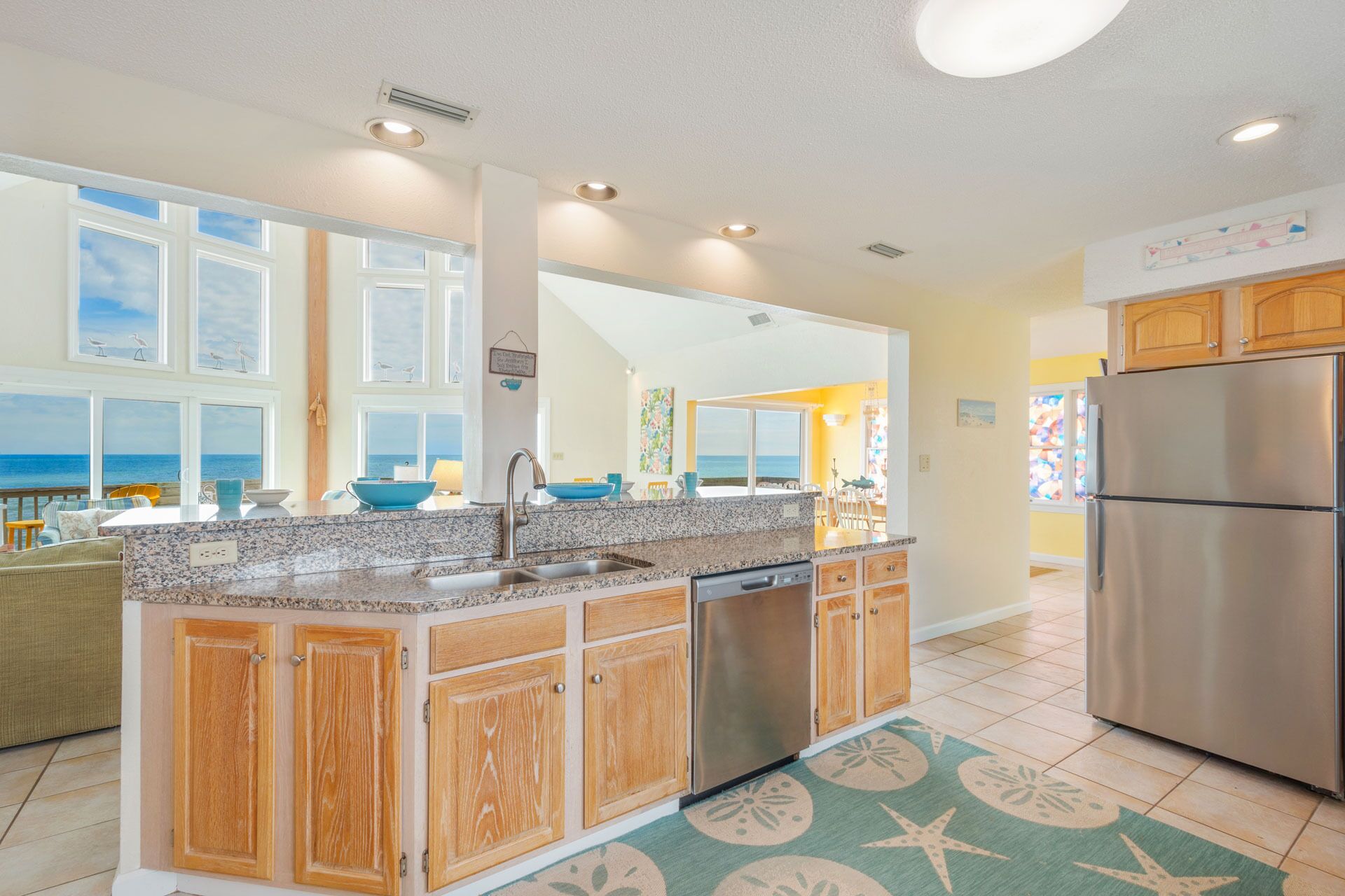 A kitchen with a granite countertop, wooden cabinets, a stainless steel refrigerator, and a view of the ocean through large windows.