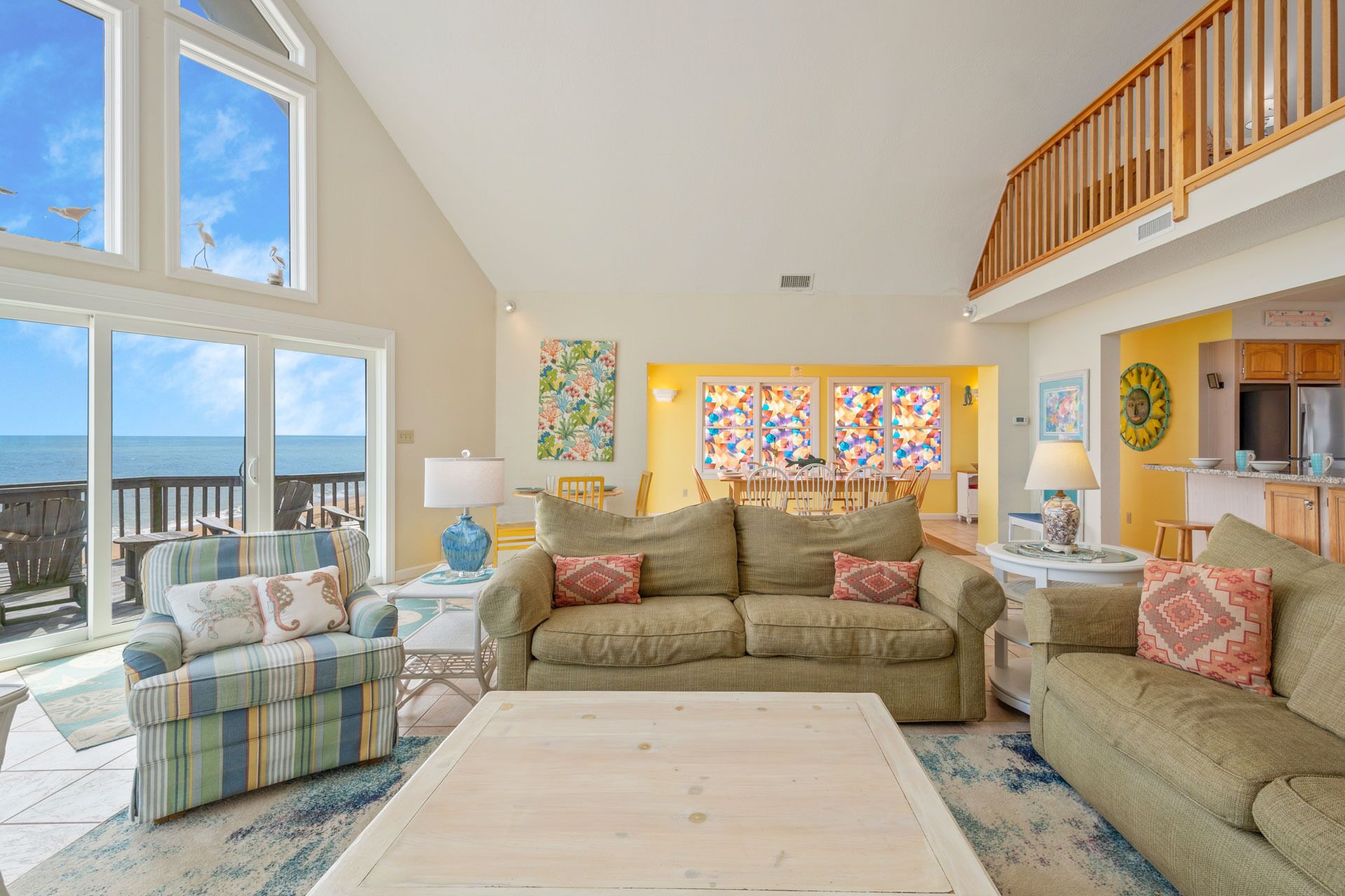 A spacious living room with a view of the ocean, a dining area with a yellow wall, and a kitchen with wooden cabinets.