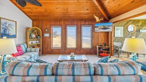 A cozy living room with a wooden ceiling, a sofa with striped cushions, a coffee table, and a view of the beach through large windows. - Cozy Oceanfront Cottage w/ Books, Games, TVs, Stunning Views. Right on the Beach (Rodanthe)