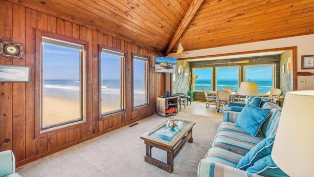 A wooden interior with large windows overlooking a beach, a sofa, a coffee table, and a TV. - Cozy Oceanfront Cottage w/ Books, Games, TVs, Stunning Views. Right on the Beach (Rodanthe)