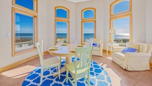 A spacious room with a round dining table, four chairs, a sofa, and a small side table. Large windows with wooden frames offer a view of the ocean.