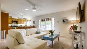 A living room with a sofa, coffee table, and a wall-mounted TV. There is a kitchen in the background with wooden cabinets and a microwave. - Driftwood your perfect beach getaway! (La Jolla)