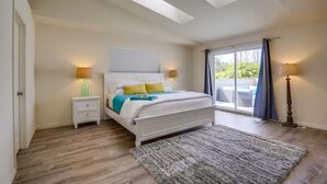 A bedroom with a skylight, a bed with a white headboard, a nightstand, a lamp, and a view through a window. - Driftwood your perfect beach getaway! (La Jolla)
