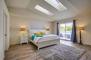 A bedroom with a skylight, a bed with a white headboard, a nightstand, a lamp, and a view through a window.