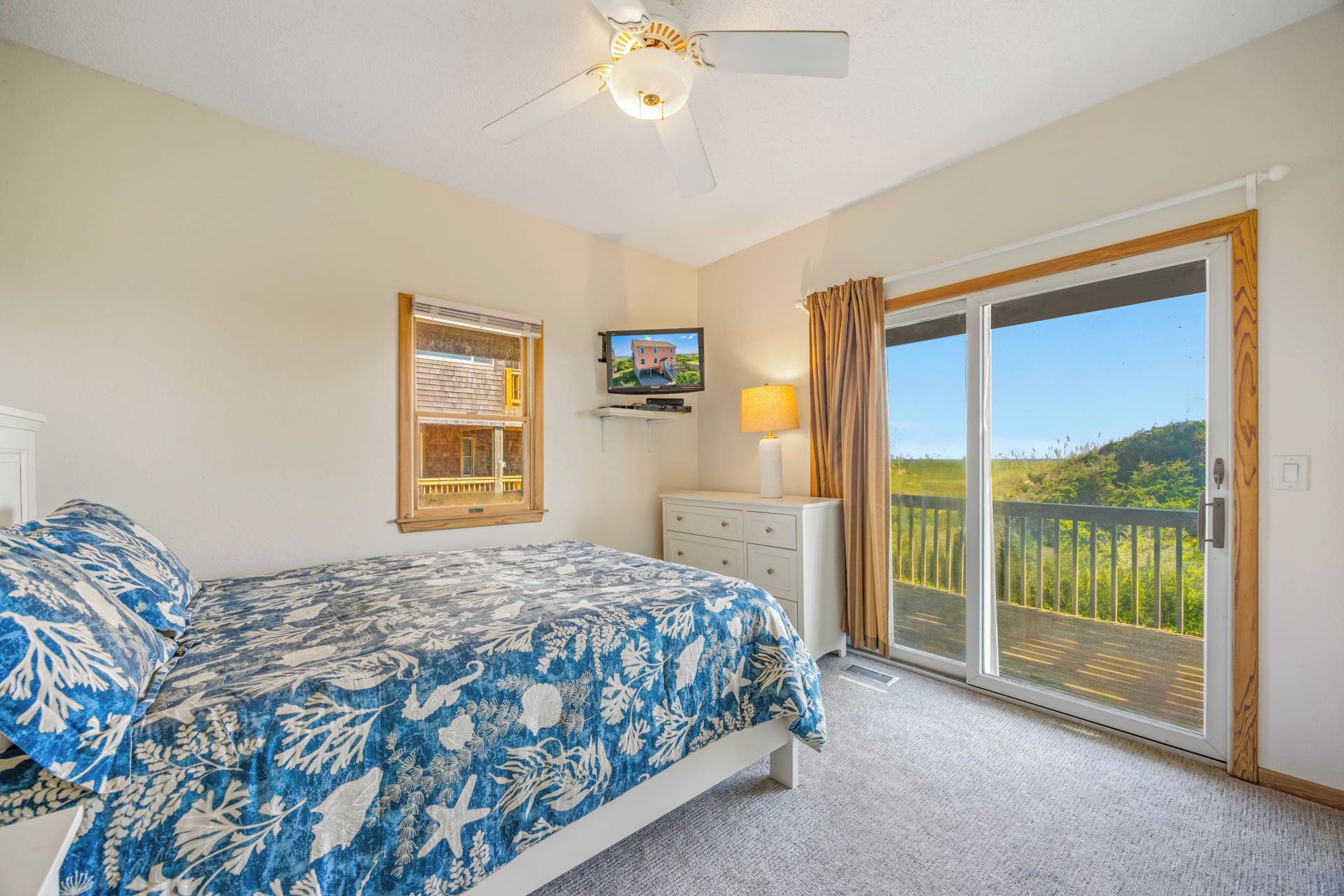 A bedroom with a bed, a television, a window with blinds, a ceiling fan, and a view of greenery through a sliding door.