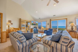 A spacious living room with striped sofas, a glass coffee table, and a view of the ocean through large windows. - Oceanfront Home in Avon with Hot Tub, Community Pool and Tennis/Pickleball (Avon)