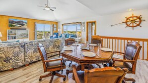 Un comedor con una mesa de madera dispuesta para cuatro personas, rodeada de sillas de cuero. Un amplio ventanal ofrece vistas a la playa.