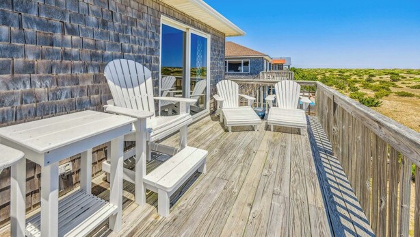 Terraza de madera con sillas y mesas Adirondack blancas, puerta de vidrio que da a un área cubierta y vista a la playa.