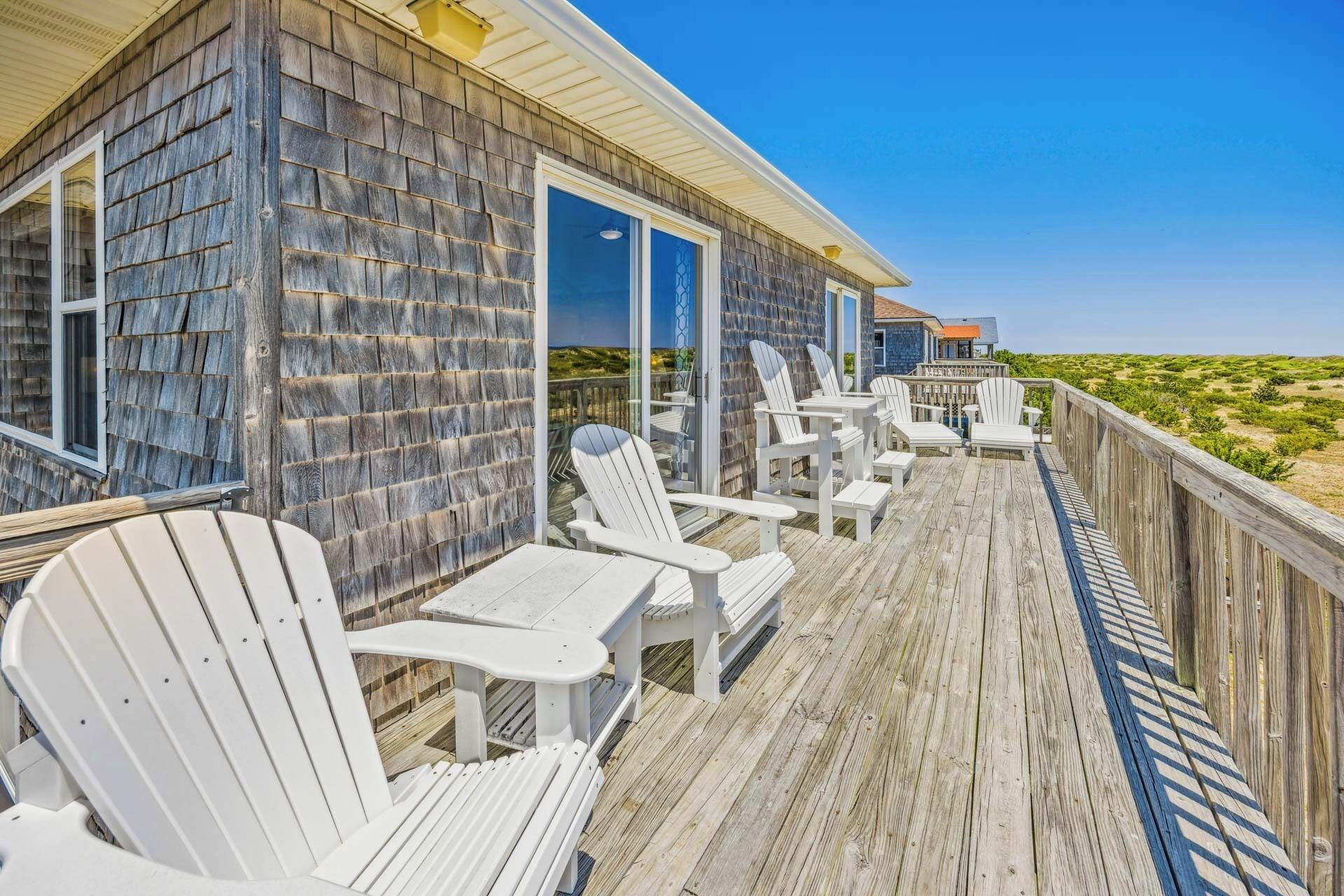 A wooden deck with white Adirondack chairs and a table, overlooking a beach.