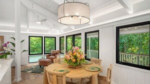 A dining area with a round wooden table, chairs, a central floral arrangement, and large windows offering a view of greenery.