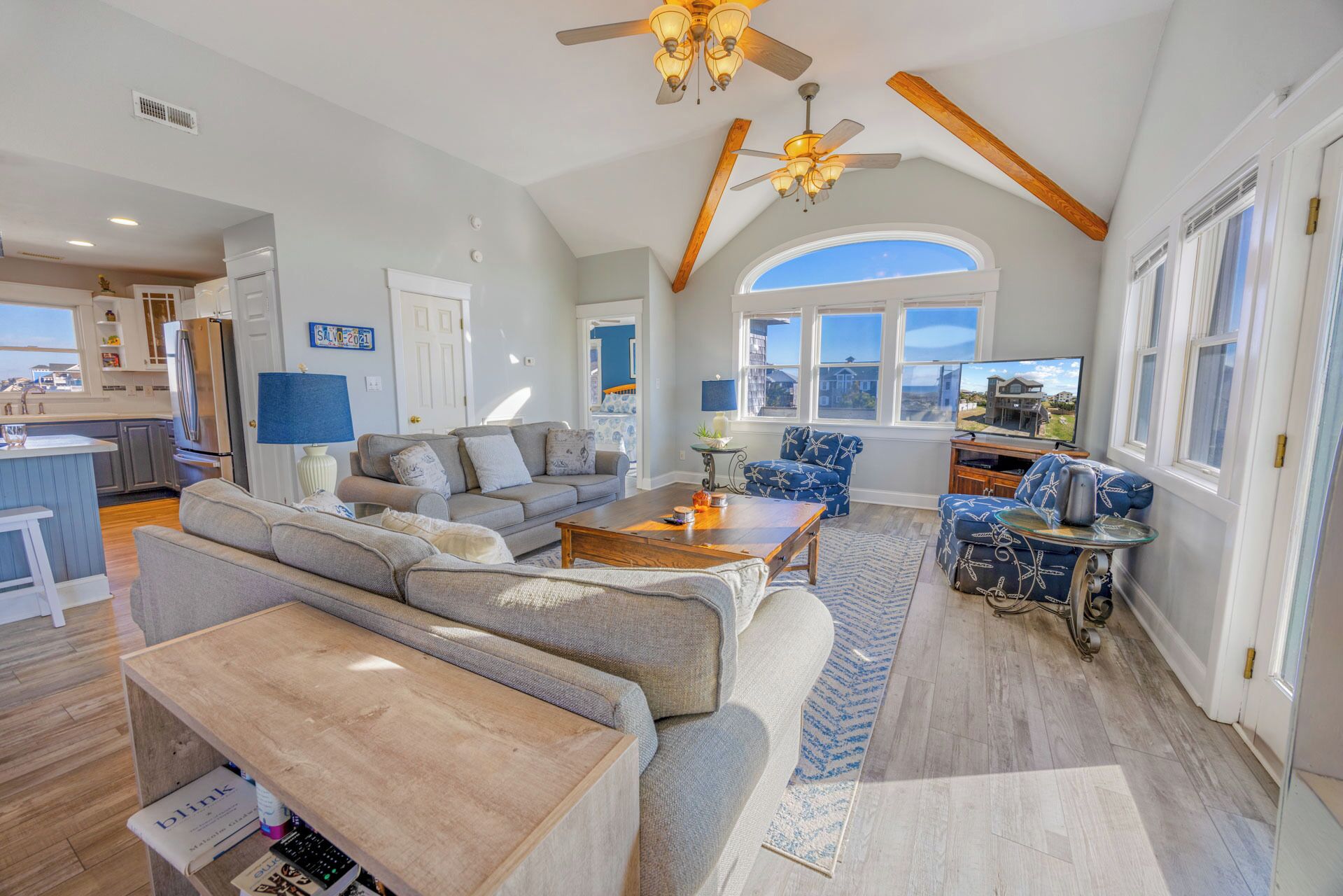 A spacious living area with a large sofa, a coffee table, and a television. There is a kitchen area with a refrigerator and a dining table. The room has wooden beams on the ceiling and large windows.