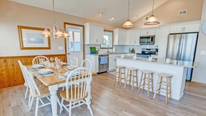 A modern kitchen with white cabinetry, a central island with stools, and stainless steel appliances. - Semi-Oceanfront Home w/ Private Pool, Hot Tub, Basketball. Watch the Waves Crest (Waves)