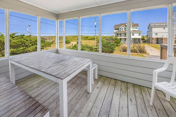 A covered outdoor area with a wooden table and chairs, large windows offering views of houses and greenery. - Semi-Oceanfront Rodanthe Home with Private Pool, Hot Tub, and Screened Porch (Rodanthe)
