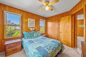 A bedroom with wooden walls, a bed with patterned bedding, a ceiling fan, and a window with a view of trees and buildings. - Oceanfront Home with Direct Beach Access, Community Pool and Tennis/Pickleball (Avon)