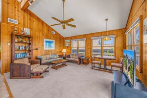 A wooden-paneled room with a ceiling fan, a sofa set, a coffee table, a dining table with chairs, and a bookshelf.