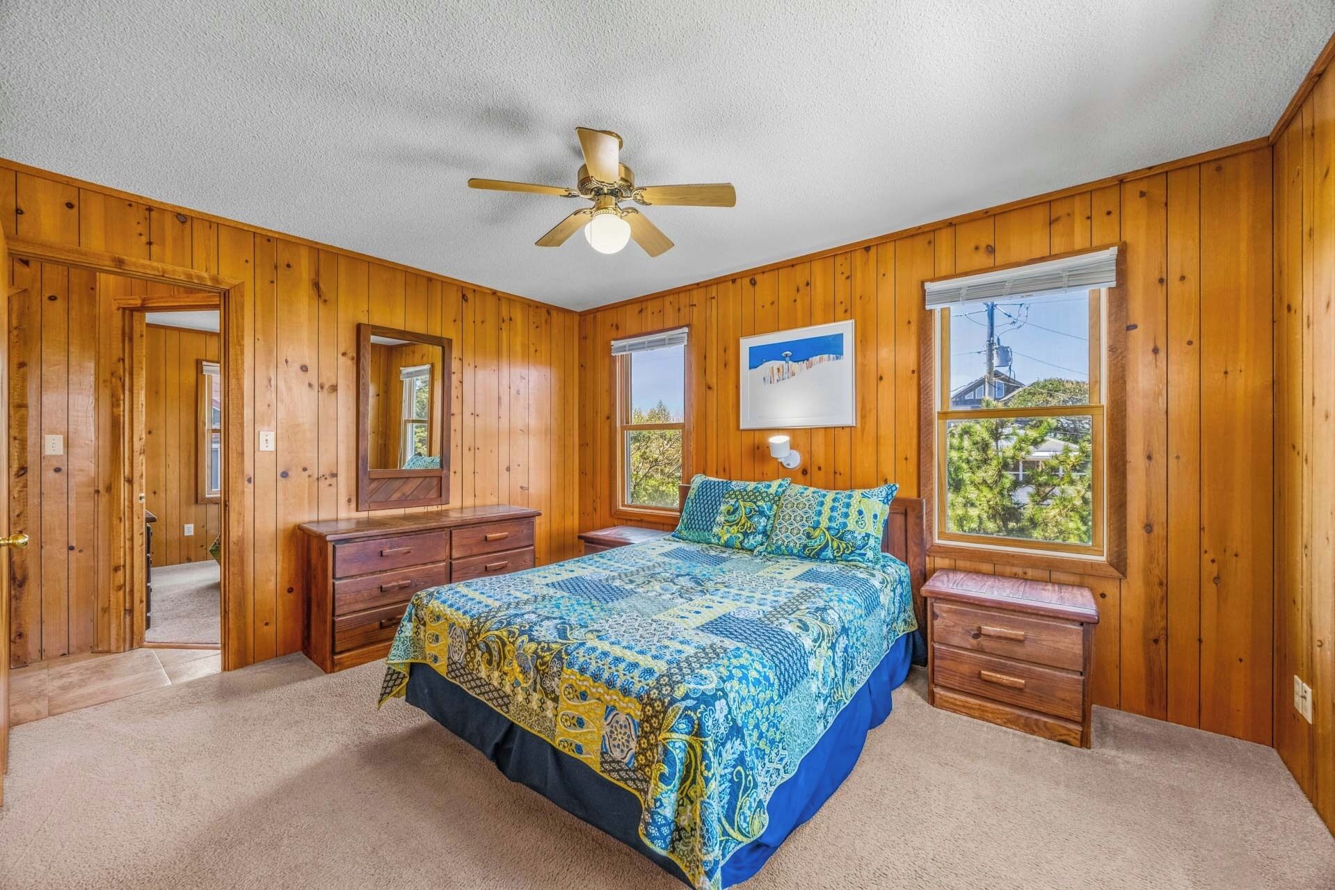 A bedroom with wooden walls, a bed with patterned bedding, a ceiling fan, and two windows with views.