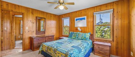A bedroom with wooden walls, a bed with patterned bedding, a ceiling fan, and two windows with views.