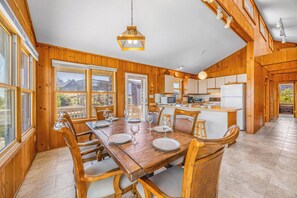 A wooden interior with a dining table set for four, kitchen area with appliances, and large windows.