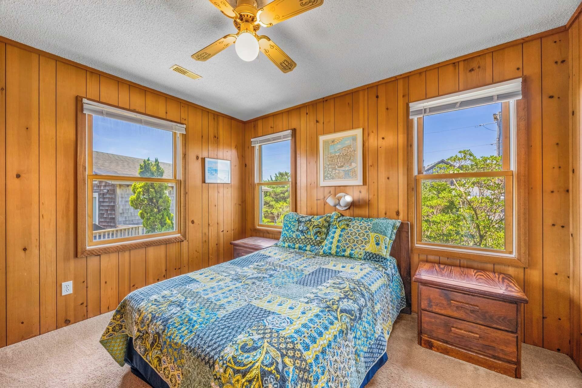 Une chambre avec des murs en bois, un lit avec une literie à motifs, une table de chevet et un ventilateur de plafond.