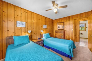 A room with two beds, wooden walls, a ceiling fan, and a door leading to another room.