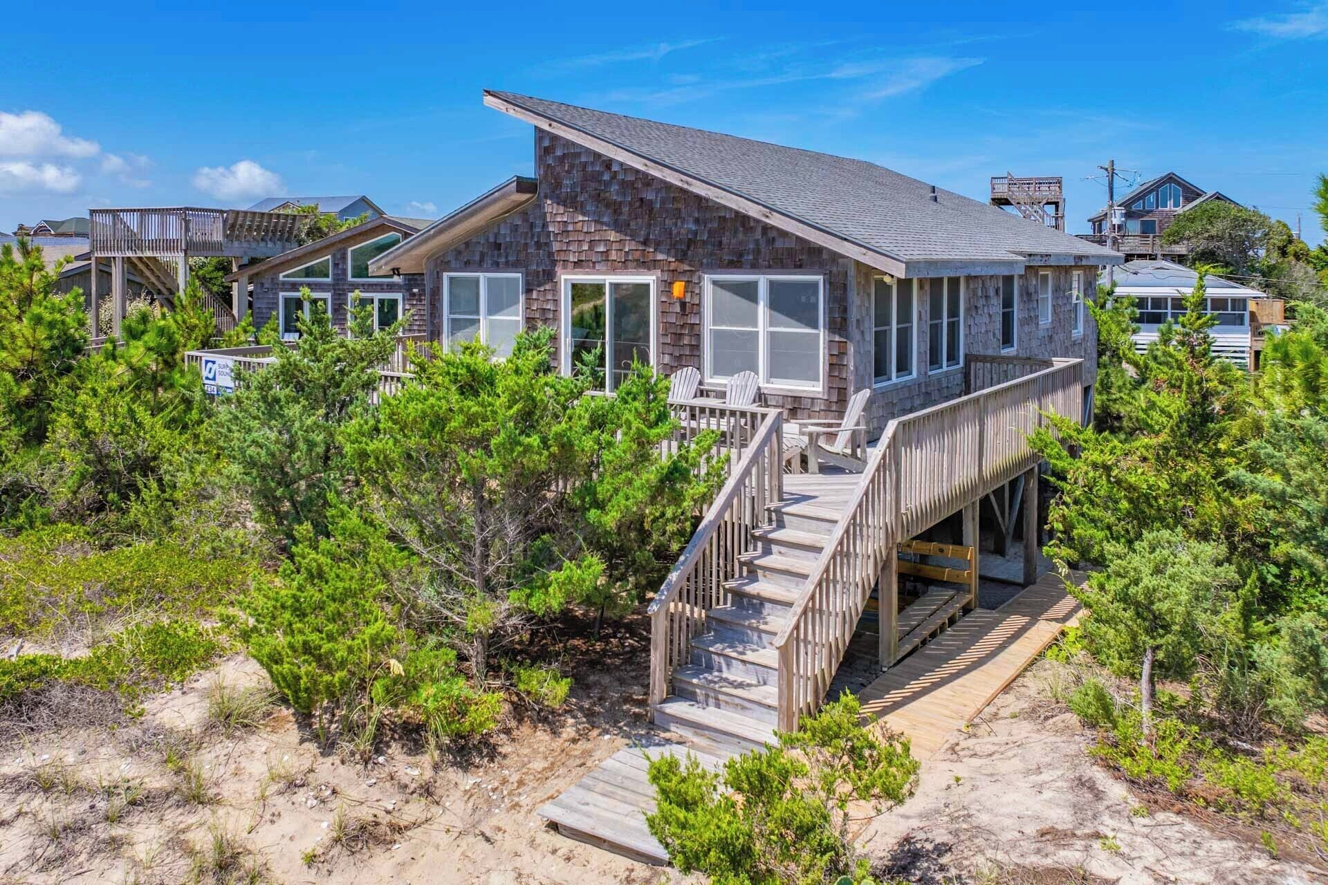 A beachfront property with a wooden deck, multiple windows, and a staircase leading to a covered area.
