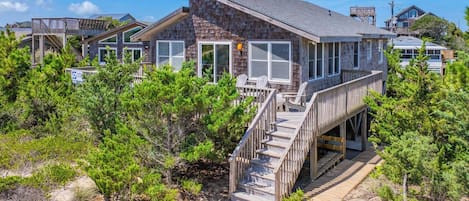 A beachfront property with a wooden deck, multiple windows, and a staircase leading to a covered area.