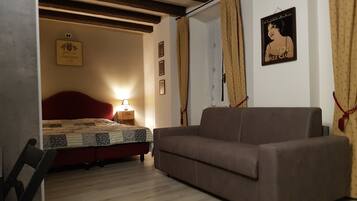 A bedroom with a bed, a sofa, a nightstand, and a wall with framed pictures.