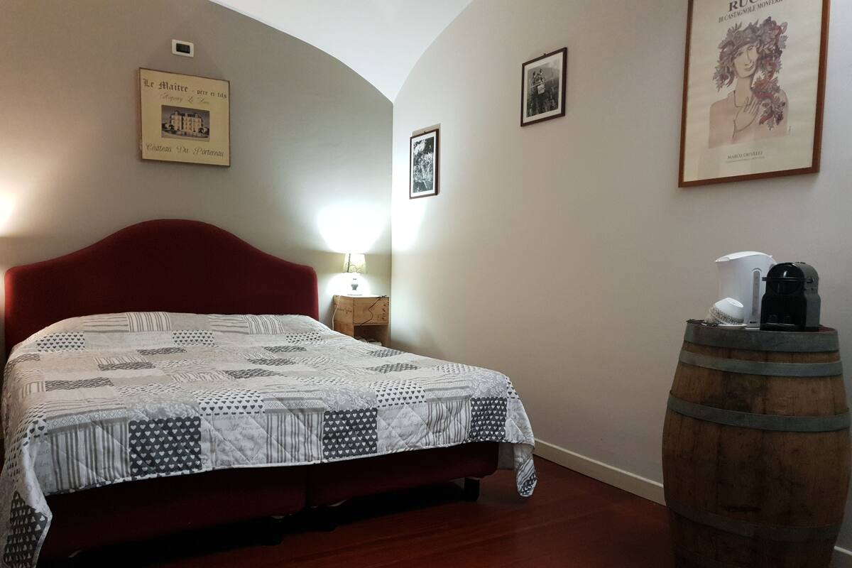 A bedroom with a red headboard, a quilted bedspread, a wooden barrel, and framed pictures on the wall.