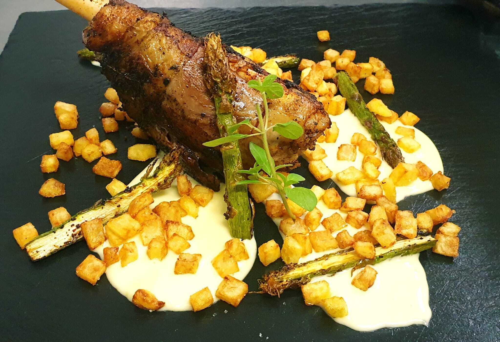 Een gebraden lamsbout met geroosterde aardappelen en asperges, gepresenteerd op een zwarte leisteenplaat.