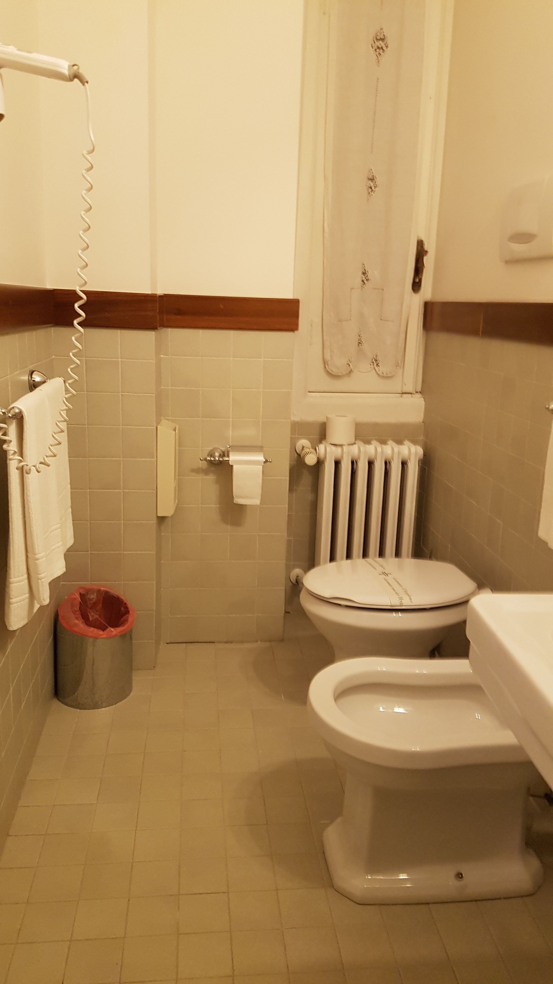 Un baño con dos inodoros, un radiador, un perchero para toallas y un cubo de basura.