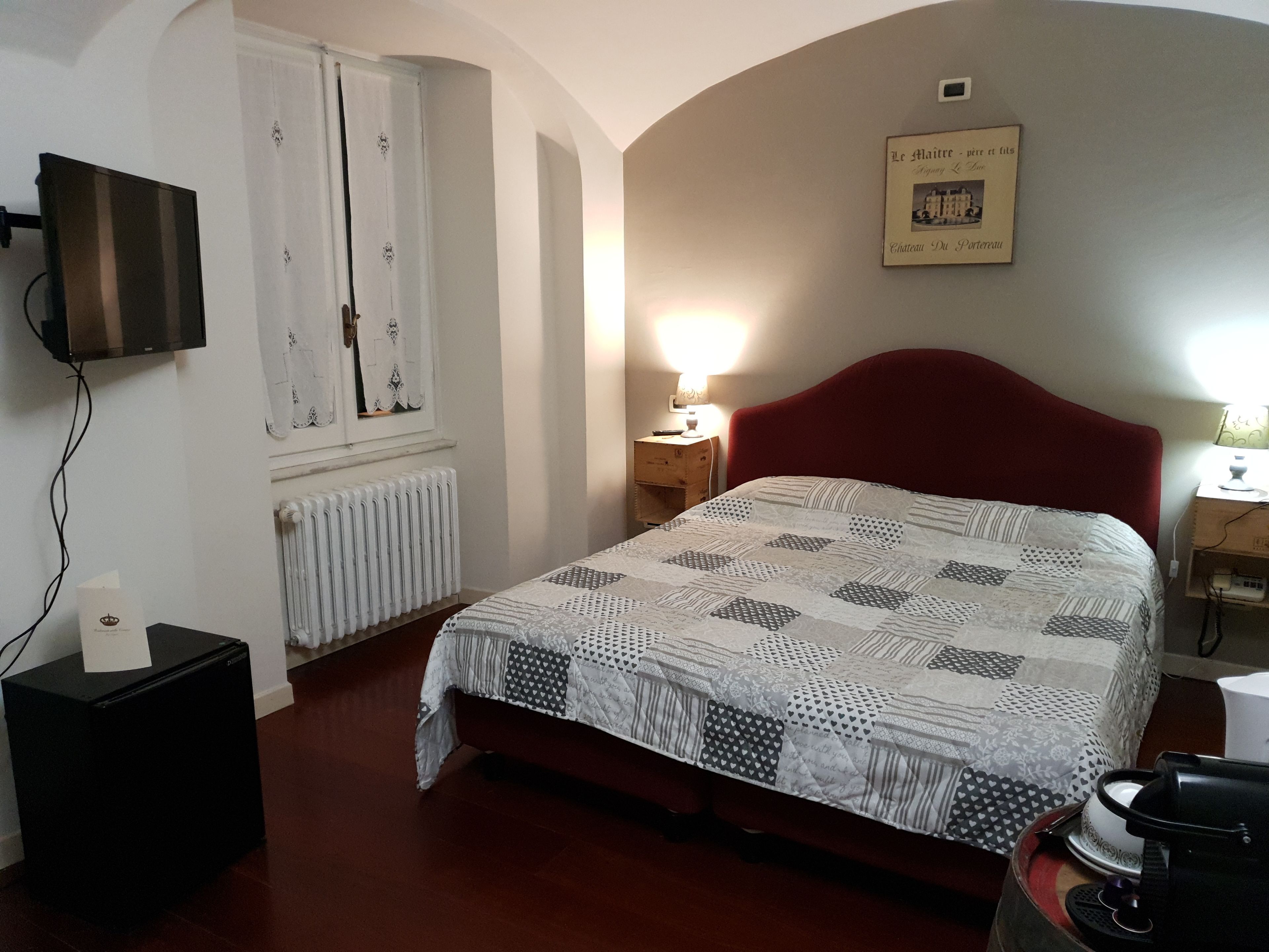 A bedroom with a red headboard, a quilted bedspread, a radiator, a television mounted on the wall, and a framed picture on the wall.