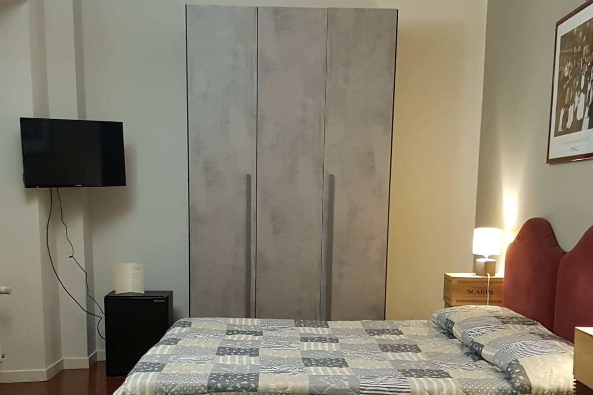 A bedroom with a large bed, a flat-screen TV mounted on the wall, a small bedside table with a lamp, and a framed picture on the wall.
