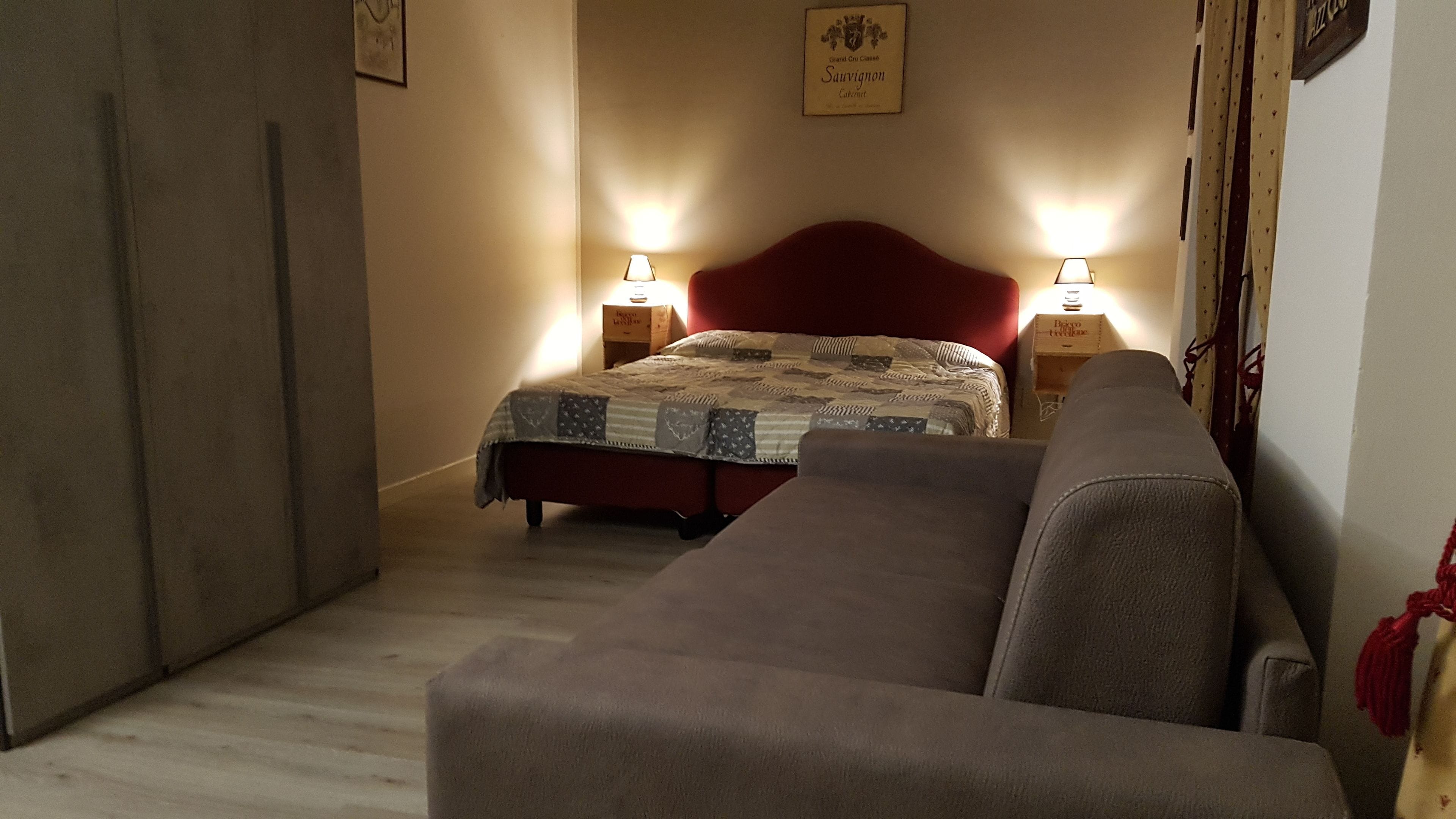 Un dormitorio con cama, dos mesitas de noche con lámparas, cabecero rojo y sofá gris.
