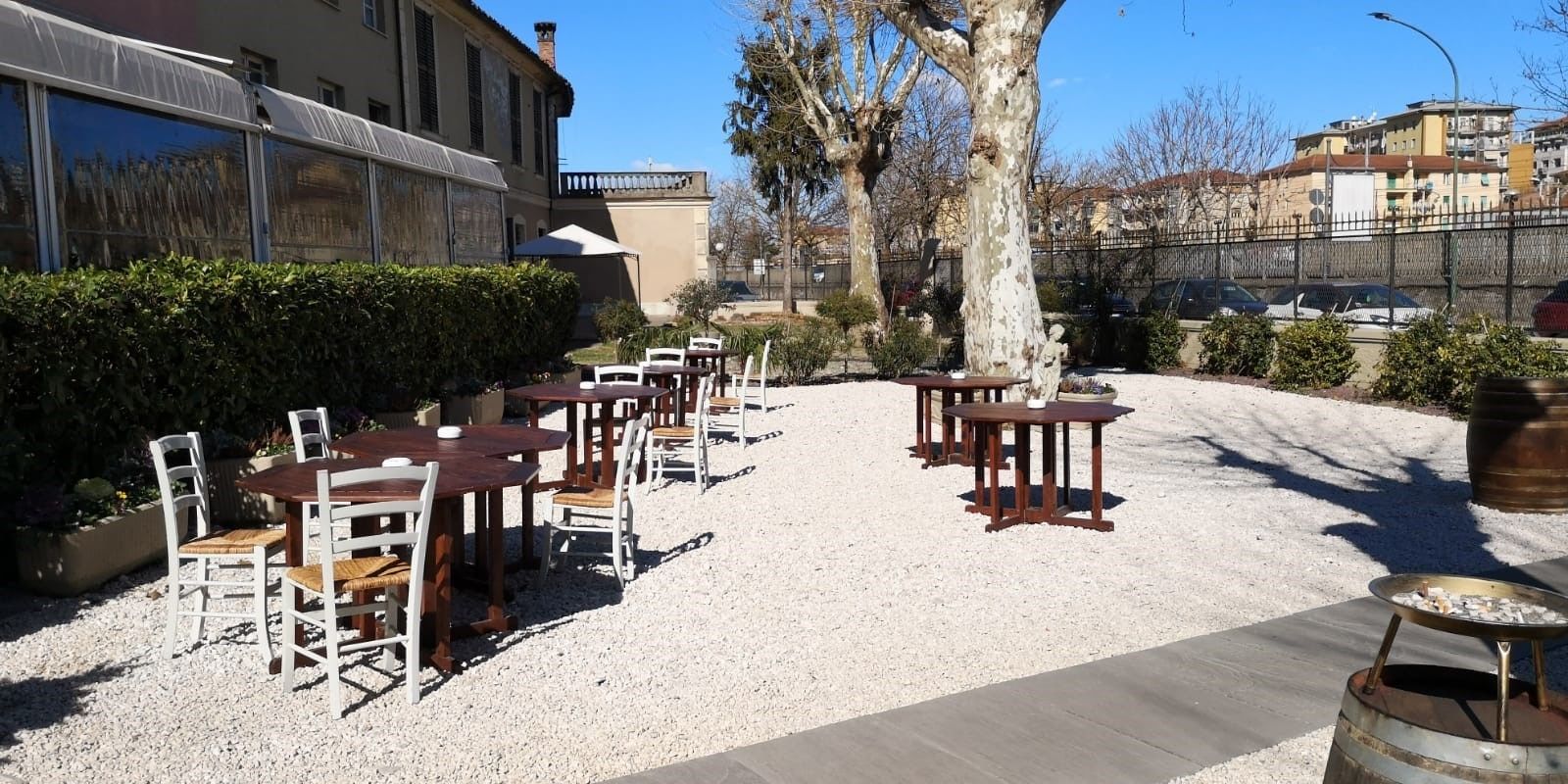 An outdoor seating area with tables and chairs, surrounded by gravel and trees, with buildings in the background.