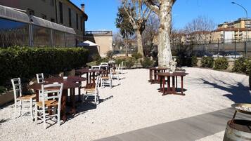 An outdoor seating area with tables and chairs, surrounded by gravel and trees, with buildings in the background.