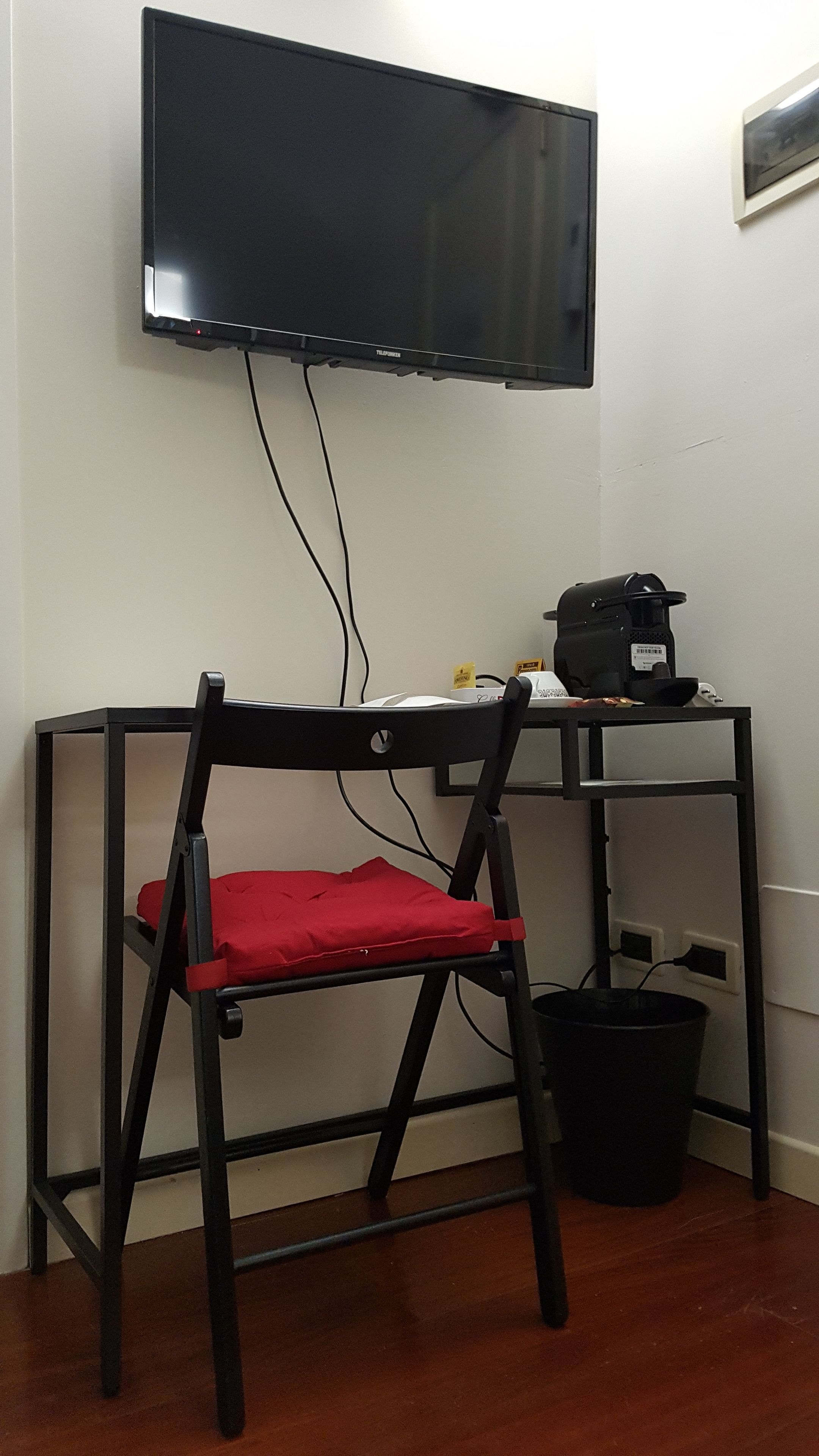 Una habitación con un escritorio de metal negro, una silla con acolchado rojo, un cubo negro y un televisor montado en la pared.