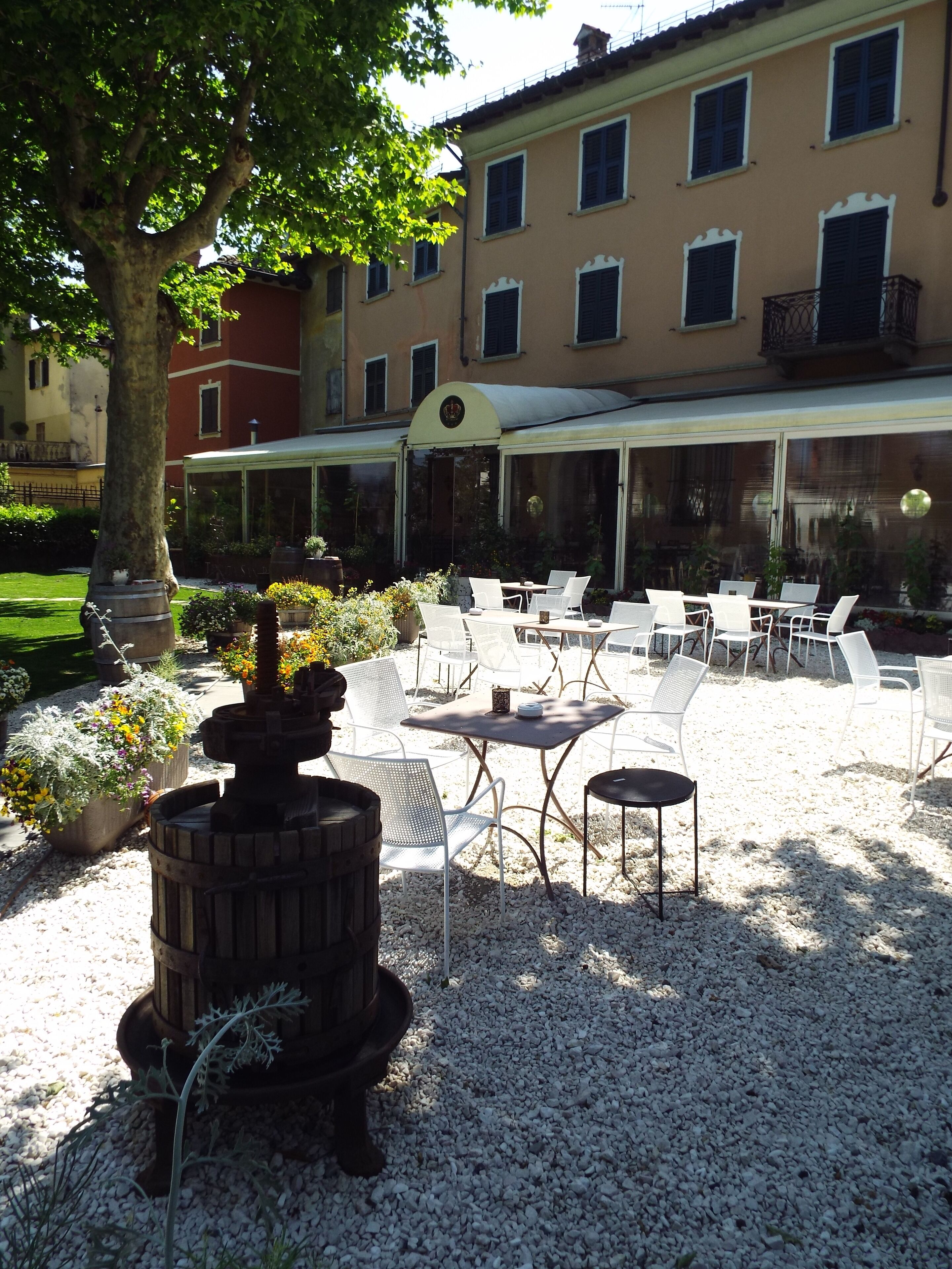 An outdoor patio with white metal chairs and tables, a decorative fire pit, and a building with multiple windows and a covered area in the background.