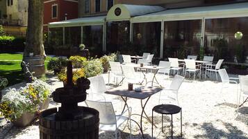 An outdoor patio with white metal chairs and tables, a decorative fire pit, and a building with multiple windows and a covered area in the background.