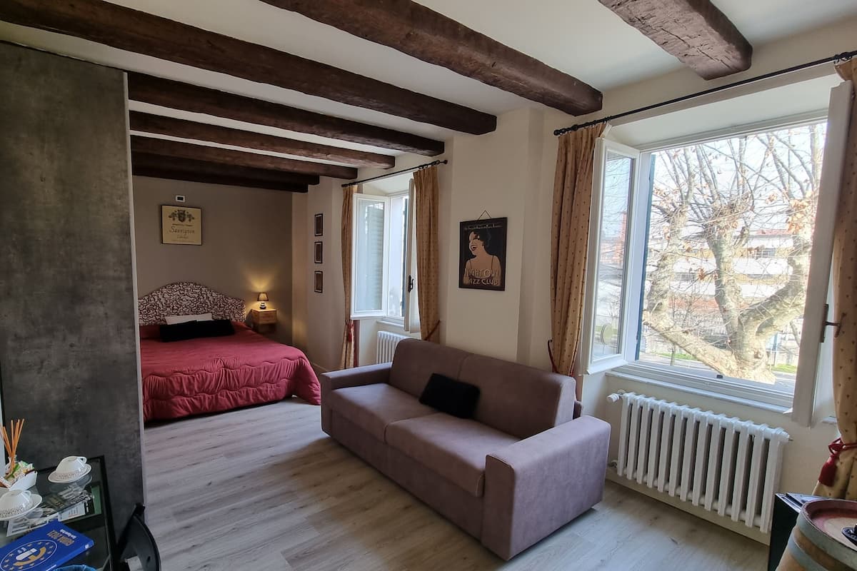 A room with a bed, a sofa, a window with curtains, a radiator, and a framed picture on the wall.