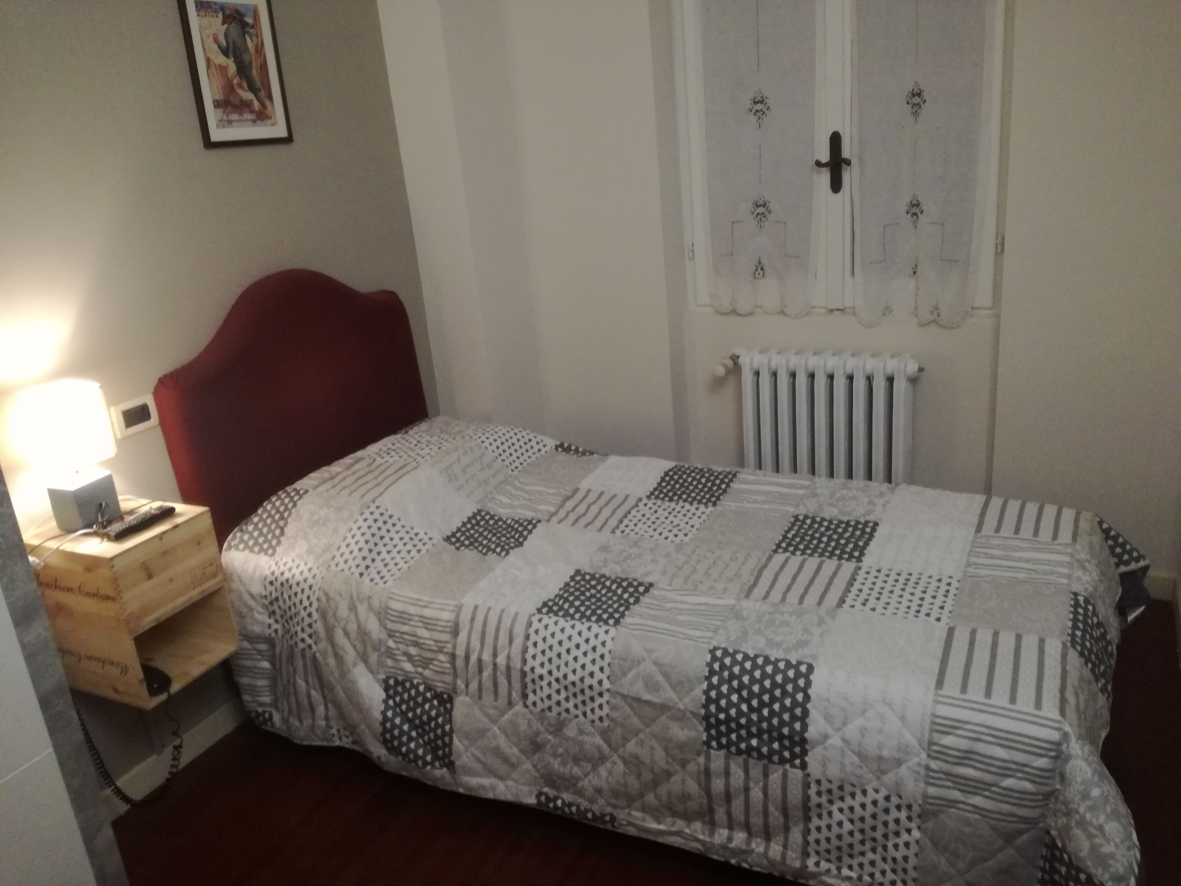 Una camera piccola con un letto singolo, un comodino, una testiera rossa, un termosifone a parete e un quadro incorniciato.