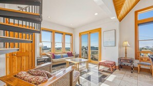 A spacious living room with a high ceiling, large windows, a staircase, and a view of the outside. - Semi-Oceanfront Hatteras Home with Private Pool, Hot Tub, Game Room and Wet Bar (Hatteras)